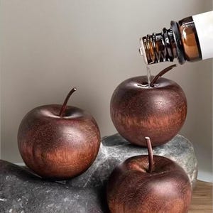 May include: Three wooden apple sculptures with stems rest on gray stones. A brown glass bottle is pouring liquid onto one of the apples. The apples have a dark brown color with a wood grain texture.