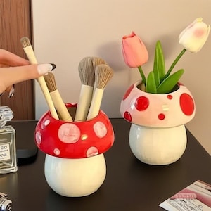 Può includere: Due contenitori in ceramica a forma di fungo. Uno è rosso con pois bianchi, contenente pennelli per il trucco. L'altro è rosa con pois rossi, contenente tulipani. Entrambi hanno basi bianche e poggiano su una superficie scura.