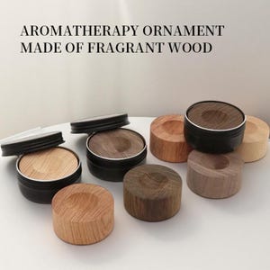 Op de afbeelding: Aromatherapie-ornamenten gemaakt van geurig hout. De afbeelding toont verschillende kleine, ronde houten aromatherapie-ornamenten. Sommige zitten in zwarte metalen blikken met deksels, terwijl andere op zichzelf staande houten stukken zijn in verschillende tinten bruin en beige.
