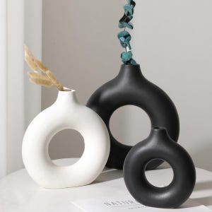 May include: Three modern ceramic vases in a minimalist style. The vases are in white and black, with a circular design. One vase holds dried beige grass, and another holds green leaves. The vases are on a white surface.