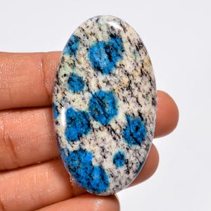 May include: Oval-shaped K2 stone, featuring a speckled white and gray base with vibrant blue orb-like patterns. The polished gemstone is held in a hand, showcasing its natural beauty and unique color variations.