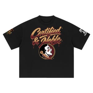 Può includere: Una t-shirt nera a maniche corte con la scritta Certified & Noble in un carattere dorato e bordeaux gocciolante sopra un logo circolare raffigurante il profilo di una persona con pittura facciale e una piuma sulle maniche.