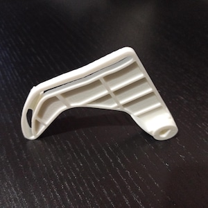 May include: A white plastic component with a curved design and rectangular openings. The object has a hole on one end and appears to be part of a mechanism. It is set against a dark surface, possibly a table.
