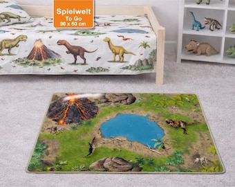 May include: A dinosaur-themed play mat with a volcano, lake, and various dinosaur illustrations. The mat is rectangular and features a detailed landscape design. A bed with a matching dinosaur-themed blanket and pillow is in the background. The text "Spielwelt To Go 90 x 60 cm" is visible.