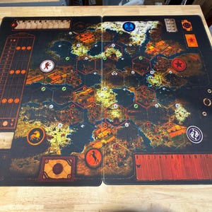 Può includere: Tappetino per giochi da tavolo in due pezzi con sfondo scuro e design a mappa. Il tappetino presenta varie icone, tra cui un orso, un cavallo e una stella. La tavolozza dei colori include sfumature di marrone, arancione e giallo, con una griglia di forme esagonali.