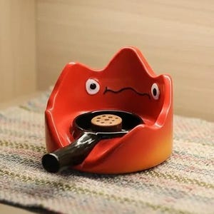 May include: A whimsical, red ceramic ashtray shaped like a flame, with a small black pan inside holding a cookie. The ashtray has two white eyes and a smiling mouth. The handle of the pan is black.