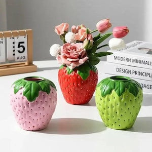 May include: Three ceramic strawberry-shaped vases in pink, red, and green. The red vase holds pink and white flowers. A wooden calendar displays "1" and "5" with "AUG" below. Books with "MODERN MINIMALISM" and "DESIGN PRINCIPLES" are in the background.