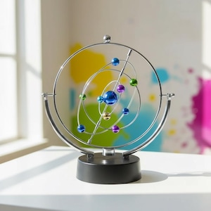 May include: A kinetic art sculpture featuring a silver-colored metal frame with orbiting spheres in various colors, including blue, green, purple, and gold. The sculpture is mounted on a black base and is designed to demonstrate the principles of orbital motion.