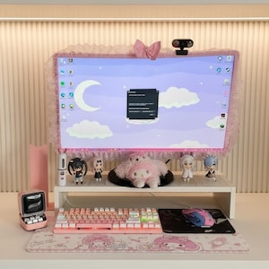Pode incluir: Uma configuração de computador com tema rosa com um monitor com moldura de renda e um laço. Um teclado branco, mouse e mousepad com personagens de desenhos animados são visíveis. Inclui estatuetas e um pequeno alto-falante rosa.