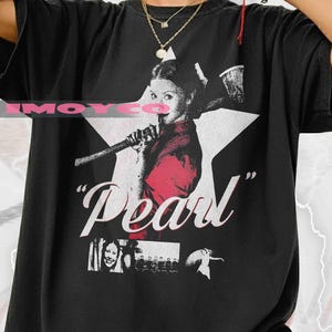 May include: Black graphic t-shirt featuring a stylized image of a person holding a microphone, set against a white star. The word "Pearl" is printed in white, with additional small images below. The shirt has a relaxed fit.