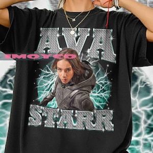 Può includere: T-shirt nero con il nome "AVA STARR" in un carattere argentato e strutturato. La maglietta mostra un ritratto di una donna in abito nero, su uno sfondo di fulmini verdi. Un capo casual.