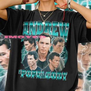 Könnte beinhalten: Schwarzes T-Shirt mit einem türkis-weißen Aufdruck. Der Aufdruck zeigt den Namen "ANDREW" und den Satz "POPE CODY", zusammen mit mehreren Bildern eines Männergesichts und einem Blitzmuster.
