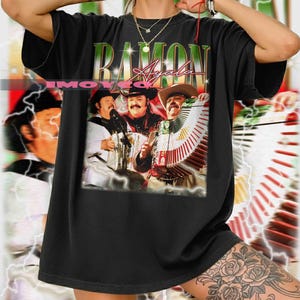 May include: Black t-shirt featuring a graphic of a musical group with the text "RAMON Ayala" in green and pink. The image shows three men playing accordions and wearing cowboy hats. The shirt has a vintage, distressed look.