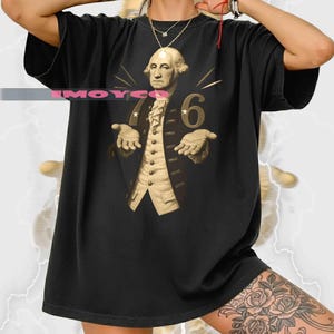 Op de afbeelding: Zwart T-shirt met een afbeelding van George Washington en het nummer 6. Het ontwerp toont Washington in formele kleding met uitgestrekte handen. Het shirt is donker van kleur.