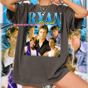 May include: Dark gray t-shirt featuring a collage of images and the text "RYAN Atwood" in a blue and white design. The shirt has a vintage aesthetic, with a faded look and a crew neck.