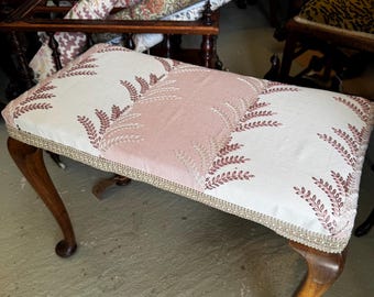 May include: A vintage upholstered bench with a rectangular seat. The seat features a pink and cream floral pattern with a gold trim. The bench has four curved wooden legs.