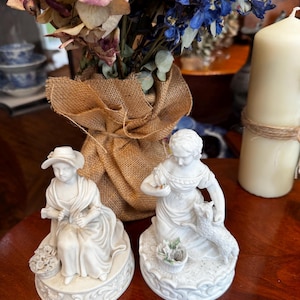 May include: Two white porcelain figurines of women. One is seated, wearing a hat and holding a basket of flowers. The other kneels, interacting with a lamb. Both are on decorative bases. A bouquet of dried flowers and a candle are in the background.