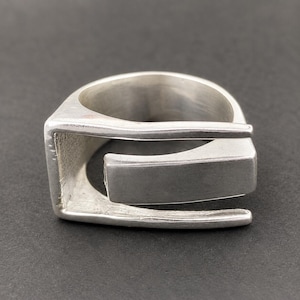 Pode incluir: Um anel de prata com um design geométrico aberto e um acabamento mate suave. A banda consiste numa estrutura angular contínua que se envolve para formar uma abertura retangular, exibindo uma estética industrial limpa com textura metálica visível.