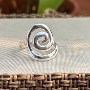 May include: A polished silver ring featuring a prominent, raised spiral design on the front. The metal has a smooth, reflective surface with a continuous, flowing shape that curves inward to form a centered coil, attached to a simple band.