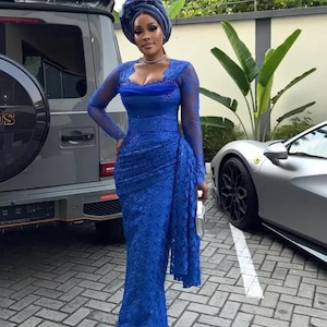 Puede incluir: Una mujer con un vestido de encaje azul real con mangas largas y un pañuelo a juego. El vestido presenta un escote corazón y un detalle lateral drapeado. Está de pie cerca de un SUV gris y un coche deportivo plateado.