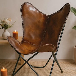 May include: A brown leather butterfly chair with a black metal frame. The chair has a curved back and seat, with visible stitching along the edges. The chair is set against a neutral background, with candles and a vase of flowers nearby.