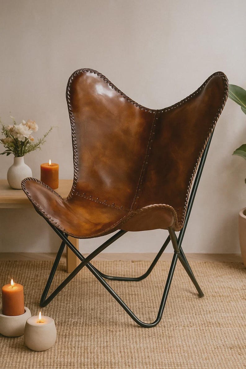 May include: A brown leather butterfly chair with a black metal frame. The chair has a curved back and seat, with visible stitching along the edges. The chair is set against a neutral background, with candles and a vase of flowers nearby.