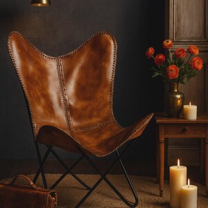 May include: A brown leather butterfly chair with a black metal frame. The chair is in a room with a jute rug, a leather satchel, and a small wooden table with a vase of orange flowers and lit candles. Three gold pendant lights hang from the ceiling.