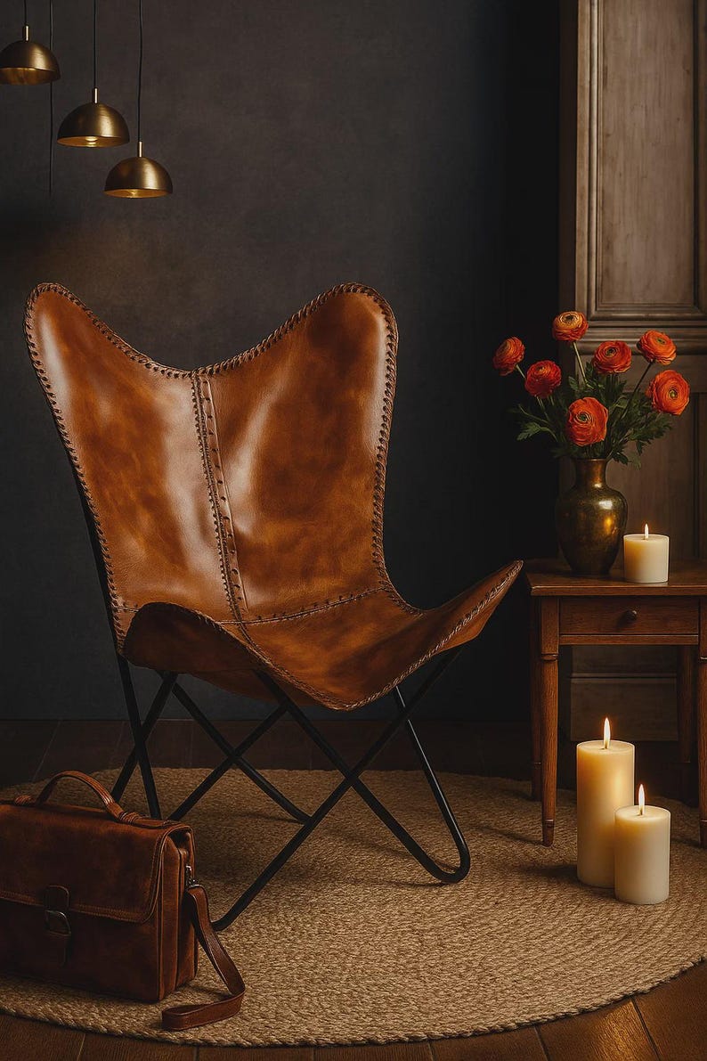 May include: A brown leather butterfly chair with a black metal frame. The chair is in a room with a jute rug, a leather satchel, and a small wooden table with a vase of orange flowers and lit candles. Three gold pendant lights hang from the ceiling.
