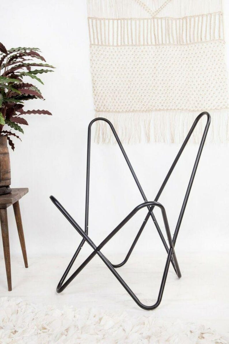 May include: Black metal butterfly chair with a minimalist design. The chair's frame is constructed from curved metal rods, creating a distinctive silhouette. The chair is suitable for indoor or outdoor use. The background includes a macrame wall hanging and a potted plant.