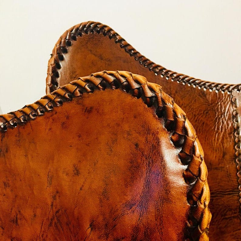 May include: Close-up of two brown leather bags with intricate braided edges. The leather has a distressed appearance with variations in color, showcasing a rustic aesthetic. The bags appear to be handmade.