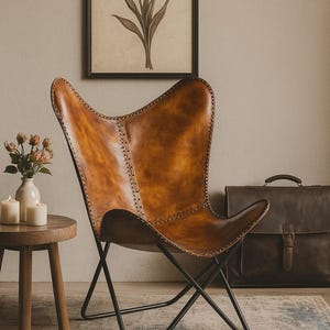 May include: A brown leather butterfly chair with a black metal frame. The chair is in a room with a framed floral print, a small wooden side table, and a leather briefcase. The chair's leather has visible stitching.