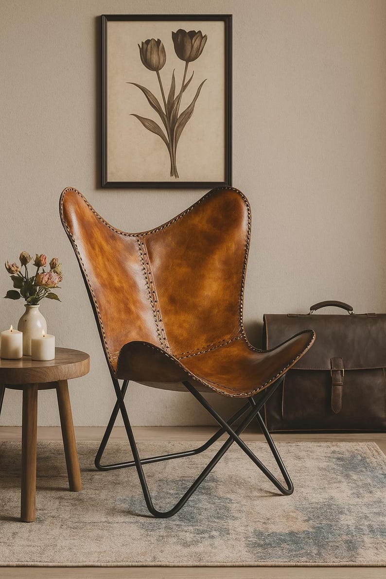 May include: A brown leather butterfly chair with a black metal frame. The chair is in a room with a framed floral print, a small wooden side table, and a leather briefcase. The chair's leather has visible stitching.