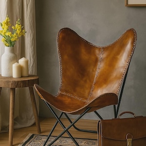 May include: A brown leather butterfly chair with a black metal frame. The chair is next to a brown leather briefcase and a small wooden table with a white vase and candles. A patterned rug is on the floor.