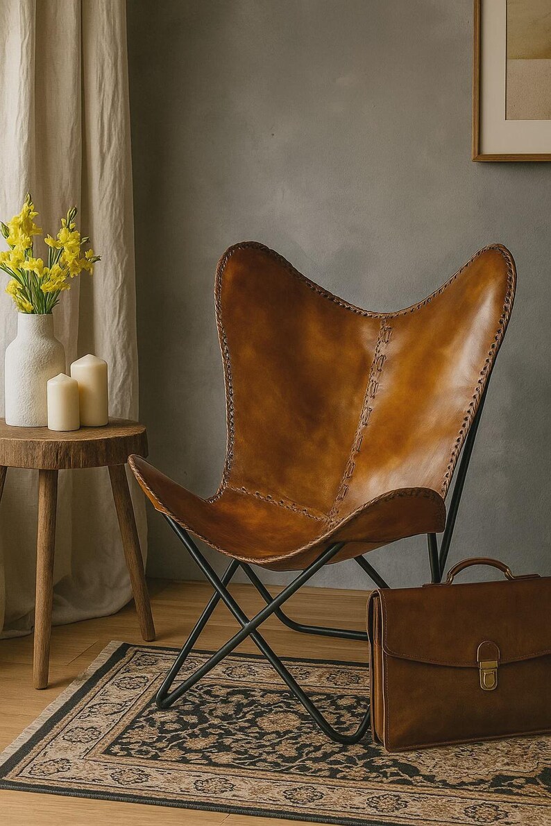 May include: A brown leather butterfly chair with a black metal frame. The chair is next to a brown leather briefcase and a small wooden table with a white vase and candles. A patterned rug is on the floor.