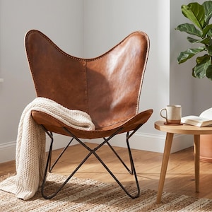 May include: A brown leather butterfly chair with a black metal frame, featuring a cream-coloured knit throw blanket draped over one side. The chair has a curved backrest and a deep seat, supported by a crossed metal base structure.