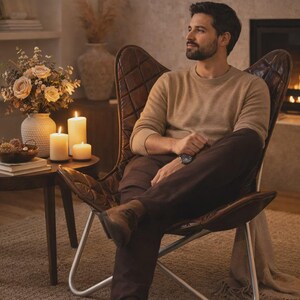May include: A brown leather butterfly chair with a diamond pattern is the focal point of a cozy living room. A person is seated in the chair, wearing a tan sweater and brown pants. Candles and flowers adorn a side table, creating a warm ambiance.
