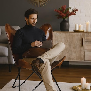 May include: A person seated in a brown leather butterfly chair, wearing a black turtleneck and tan pants. The person is holding a brown leather portfolio. The room features a gold sunburst mirror and a wooden cabinet.
