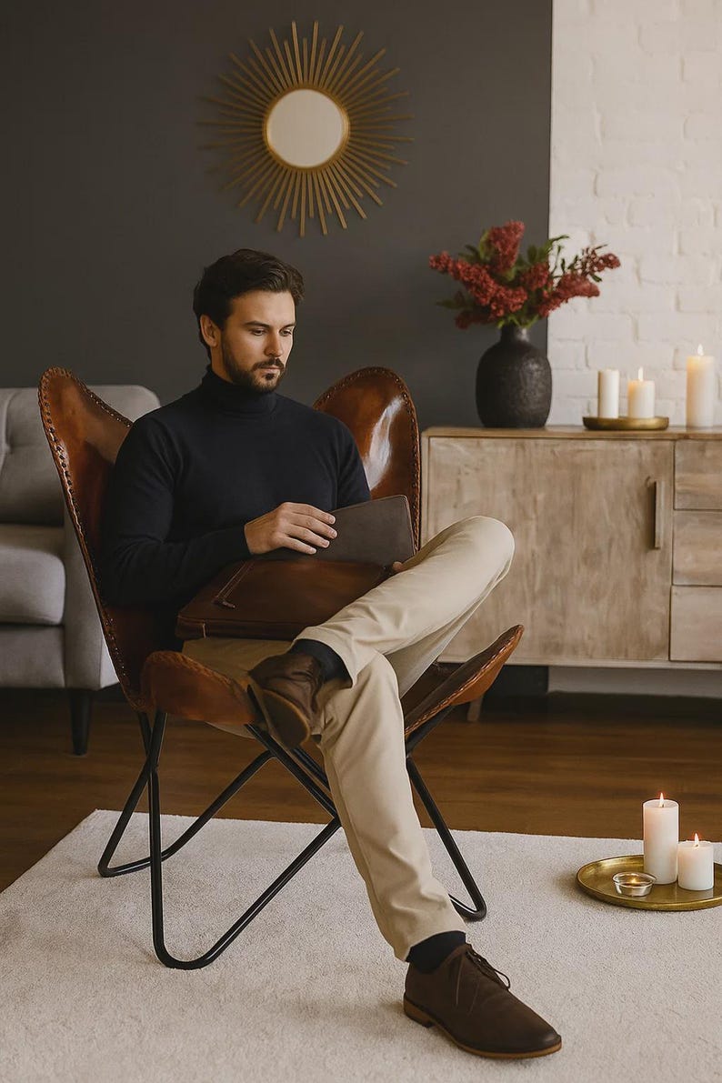 May include: A person seated in a brown leather butterfly chair, wearing a black turtleneck and tan pants. The person is holding a brown leather portfolio. The room features a gold sunburst mirror and a wooden cabinet.