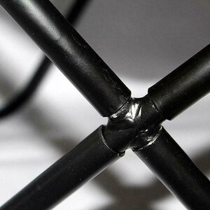 May include: Close-up of a black metal frame joint, with four cylindrical bars intersecting at a central point. The bars are welded together, creating a sturdy and industrial aesthetic. The background is blurred.