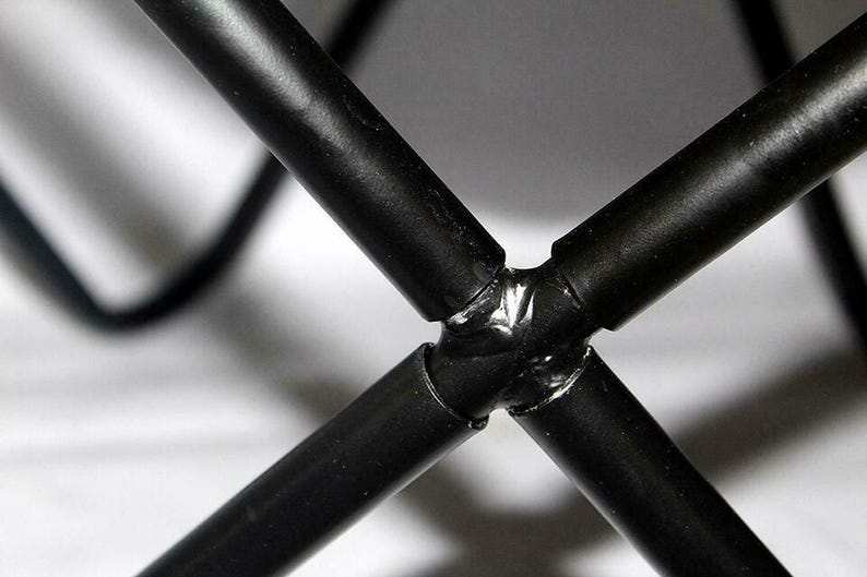 May include: Close-up of a black metal frame joint, with four cylindrical bars intersecting at a central point. The bars are welded together, creating a sturdy and industrial aesthetic. The background is blurred.