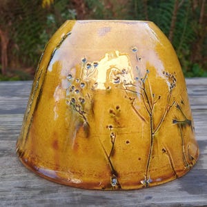 Può includere: Un vaso in ceramica fatto a mano con una base ampia e una parte superiore svasata. Il vaso è smaltato in una calda tonalità giallo dorato e presenta un design botanico in rilievo di delicati steli e capolini. Il vaso è alto circa 15 cm.