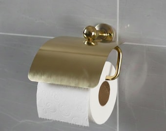 May include: A brass-toned toilet roll holder fixed to a grey tiled wall. The holder features a curved cover and a curved bar for the toilet roll. A roll of white toilet paper is partially unrolled.