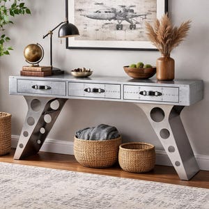 Puede incluir: Una consola de metal plateado con tres cajones y tiradores negros. La mesa tiene patas anguladas con recortes circulares. Los elementos decorativos incluyen un globo terráqueo, libros y una lámpara de escritorio negra. Cestas tejidas y una impresión enmarcada de un avión completan la decoración.
