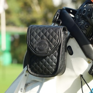May include: Black quilted leather golf accessory pouch with the text "DIALED IN" embossed on the front. The pouch is attached to a white golf bag. The background is a blurred green golf course.