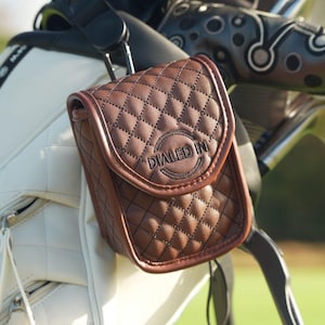 Könnte beinhalten: Eine braune, gesteppte Golf-Zubehörtasche aus Leder mit dem Text "DIALED IN" auf der Vorderseite. Die Tasche ist an einer weißen Golftasche mit einem schwarzen Riemen befestigt. Der Hintergrund ist ein grüner Golfplatz.