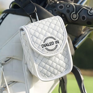 May include: White quilted golf accessory bag with the text "DIALED IN" in black. The bag is attached to a golf bag, with a green background. The bag has a flap closure and a diamond pattern.