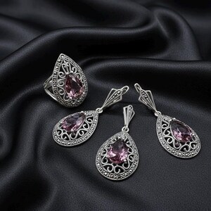May include: A silver jewellery set comprising a ring and drop earrings, each with a light purple, teardrop-shaped gemstone. The ring and earrings have an ornate filigree design with small, dark accents, set against a black satin background.