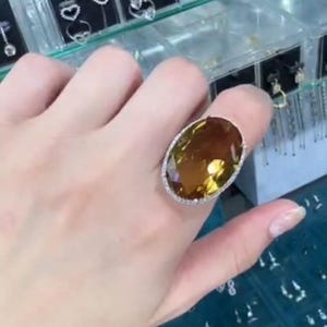 Op de afbeelding: Een ring met een grote, ovaal geslepen, barnsteenkleurige edelsteen in een zilveren zetting, omringd door een halo van kleine, heldere steentjes. De ring wordt getoond voor doorzichtige opbergdoosjes met diverse zilveren oorbellen en sieraden.