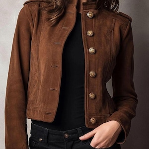 May include: A brown suede jacket with a stand-up collar and gold buttons. The jacket has a tailored fit and features shoulder epaulets. The model is wearing a black shirt and black jeans. The jacket is a classic style.