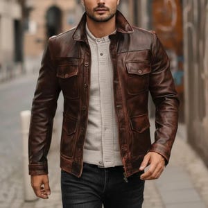 Può includere: Giacca in pelle marrone con quattro tasche frontali e colletto. La giacca è indossata sopra una camicia grigio chiaro con bottoni e jeans scuri. La giacca ha un aspetto robusto e usurato.